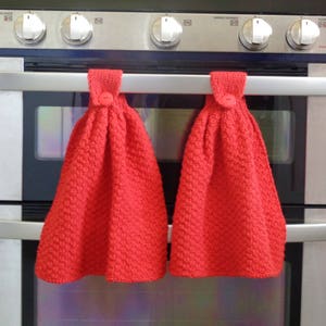 May include: Two red knitted dish towels hanging on a silver oven handle. The towels have a textured pattern and a button on the top.