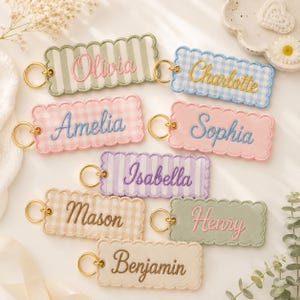 May include: A collection of rectangular embroidered keychains with scalloped edges and gold-tone metal rings. Each keychain features a unique name stitched in cursive thread, including Olivia, Charlotte, Amelia, Sophia, Isabella, Mason, Henry, and Benjamin.