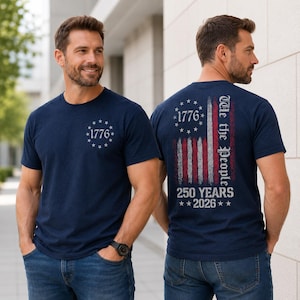 Op de afbeelding: Een marineblauw t-shirt met korte mouwen en een kleine witte afbeelding op de borst met de tekst 1776 omringd door sterren. De achterkant toont een verweerd Amerikaans vlagontwerp met de tekst 1776, We the People en 250 YEARS 2026.
