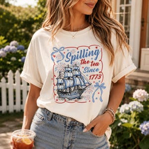 May include: A cream-colored short-sleeve t-shirt featuring a graphic of a blue sailing ship, red fireworks, and blue ribbons. The text reads Spilling the tea Since 1773. The design is enclosed in a wavy red border with small red stars.