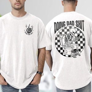 Op de afbeelding: Wit t-shirt met zwarte grafische ontwerpen. De voorkant heeft een handgebaar en de tekst "DOING DAD SHIT". De achterkant toont een skelet op een toilet, met een telefoon, met de tekst "DOING DAD SHIT" in een cirkel.