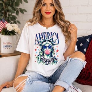May include: A white short-sleeve t-shirt featuring a graphic of the Statue of Liberty wearing star-shaped blue sunglasses and a pink bubble, with the word AMERICA printed above in blue, surrounded by small red and blue stars and a beaded necklace.