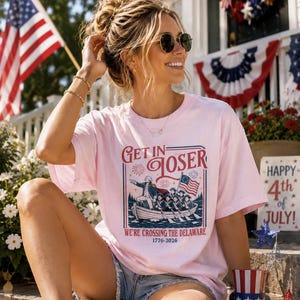 May include: A light pink short-sleeve t-shirt featuring a graphic print of George Washington in a boat with soldiers, the text "GET IN LOSER WE'RE CROSSING THE DELAWARE 1776-2026", and red, white, and blue accents including fireworks and an American flag.