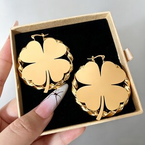 May include: A pair of gold-colored clover-shaped hoop earrings. Each earring features a four-leaf clover design within a textured, circular frame. The earrings are displayed in an open, tan-colored gift box with a black interior.