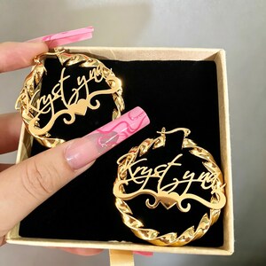 May include: A pair of gold-toned hoop earrings featuring a twisted metal design. Each earring displays the name Krystyna in a cursive script font, integrated into the centre of the hoop with a small heart detail positioned below the name.