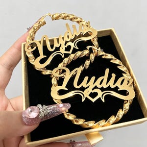 May include: Gold-colored hoop earrings with the name "Nydia" in a cursive font. The earrings have a twisted rope design and are displayed in a small box. A heart design is below the name.