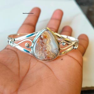 Puede incluir: Un brazalete de plata con una piedra central en forma de lágrima que presenta bandas intrincadas y arremolinadas de colores blanco, gris y marrón rojizo. La banda de metal muestra volutas decorativas y detalles en plata pulida.