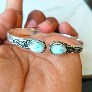 Könnte beinhalten: Ein silberner Armreif mit zwei tropfenförmigen, hellblauen Larimar-Steinen an den offenen Enden. Das Metallband zeigt filigrane Schnörkel und gedrehte Kordeldetails um jeden Stein. Der Text AmericanJewelrys ist auf dem Schmuckstück zu sehen.