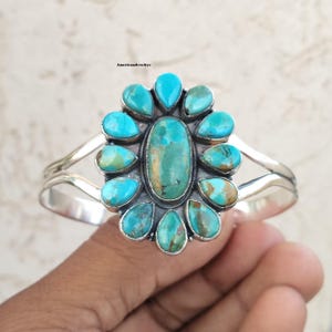 May include: A silver cuff bracelet featuring a large central oval turquoise stone surrounded by eleven smaller teardrop-shaped turquoise stones. The metal setting has a polished finish, and the text AmericanJewlrys is visible above the bracelet.