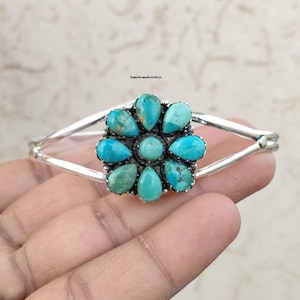 May include: A silver cuff bracelet featuring a central flower-shaped cluster of nine teardrop-shaped turquoise stones with a smooth finish. The metal band splits into two arms on each side of the floral centrepiece. The text AmericanJewlrys is visible.