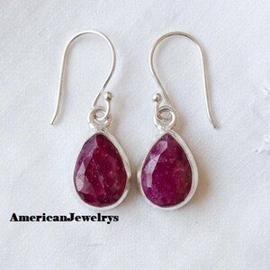 May include: A pair of silver drop earrings featuring faceted, teardrop-shaped deep pink stones. Each earring has a curved silver hook wire and a simple metal setting surrounding the stone. The text AmericanJewelrys is visible.