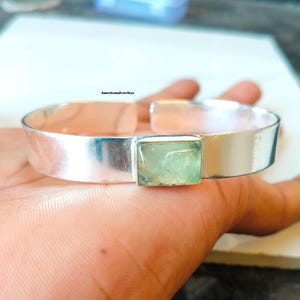 May include: A silver cuff bracelet featuring a smooth, polished band and a single rectangular, light green gemstone set in the centre. The metal has a reflective finish, and the text AmericanJewelrys is visible above the centre of the bracelet.