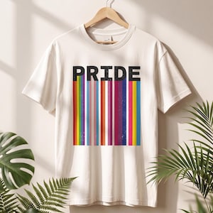 Könnte beinhalten: Ein cremefarbenes Kurzarm-T-Shirt mit dem Wort PRIDE in fetten, schwarzen Großbuchstaben. Unter dem Text befindet sich eine Reihe vertikaler Streifen in verschiedenen Farben, darunter Rot, Orange, Gelb, Grün, Blau, Lila, Pink und Weiß.