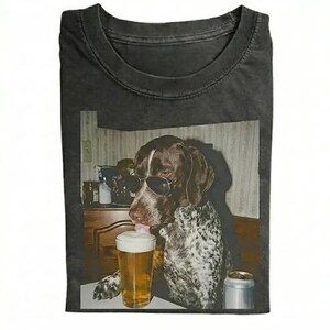 May include: A charcoal gray crew neck t-shirt featuring a graphic print of a brown and white spotted dog wearing black sunglasses. The dog is positioned next to a glass of beer with a thick white foam head and a silver aluminum can on a table.