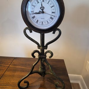 May include: A round quartz clock with a dark brown frame and a white face featuring black Roman numerals. The clock is mounted on a dark, weathered metal stand with decorative scrollwork and a tripod base, providing a stable structure.