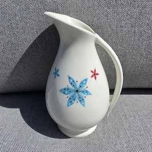May include: A white ceramic pitcher featuring a hand-painted floral pattern. The design includes a large central blue flower with dotted petals, a smaller blue flower to the left, and a single red flower to the right, all with thin black stems.