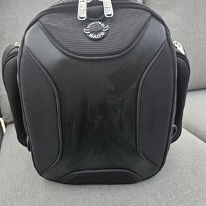 May include: A black motorcycle tail bag with a structured, molded exterior. The bag features a central Viking Bags logo, silver-toned metal zipper pulls, and side compartments. The surface has a smooth, matte finish with textured fabric accents.