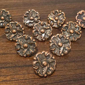 May include: A collection of ten round metal buttons featuring a raised, symmetrical floral pattern with a central dome. The buttons have a dark, oxidized copper finish with highlights on the embossed leaf-like petals and a smooth, circular edge.