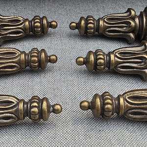 May include: A set of six antique brass-toned metal drawer pulls arranged in two columns. Each pull features a symmetrical, elongated spindle shape with intricate, repeating vertical ridges and rounded finials at both ends of the hardware.