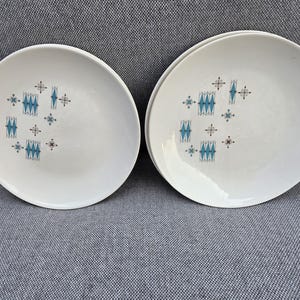 May include: Two round white ceramic plates featuring a repeating geometric pattern in shades of teal blue and dark brown. The design consists of stylized diamond shapes and small star-like accents arranged in a cluster on the surface of each plate.