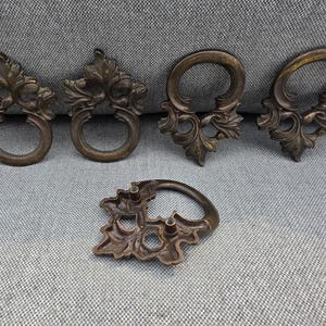 May include: Five bronze-colored metal drawer pulls featuring ornate, leaf-like decorative backplates and circular drop handles. Four pulls are displayed upright, while one is positioned flat to reveal the two mounting posts on the reverse side.
