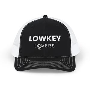 May include: Black and white trucker hat with the text "LOWKEY LOVERS" in white block letters. The hat features a white mesh back and a curved brim. A symbol of a female gender is between the words.