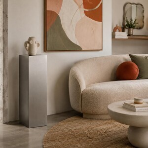 Könnte beinhalten: Ein modernes Wohnzimmer mit beigem Sofa, Kissen und rundem Couchtisch. Ein neutralfarbener Sockel trägt eine dekorative Vase. Ein abstraktes Gemälde und ein Jute-Teppich ergänzen das stilvolle Interieur.