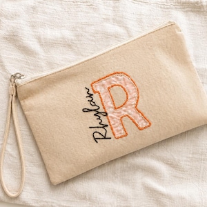 May include: A beige canvas zippered pouch featuring a wrist strap and an embroidered orange letter R. The name Rhylan is stitched in black cursive script next to the letter. The pouch has a simple rectangular shape and a metal zipper pull.