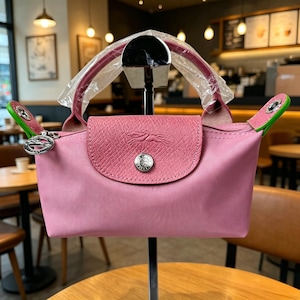 May include: A pink handbag with a flap closure and a top handle. The bag features a textured flap with a logo and a silver-tone button. The handle is burgundy, and the accents are green. The bag is displayed on a stand.