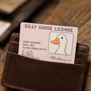 May include: A brown leather wallet holds a "Silly Goose License" card. The card features a cartoon goose illustration and text including "DOB: 4/20/69", "SEX: YES", and "CLASS: D(EEZ NUTS)". The wallet is partially open, revealing the card.