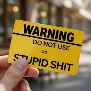 May include: A yellow rectangular sign with black text that reads "WARNING DO NOT USE on STUPID SHIT". The sign is held by a hand, with a blurred background.