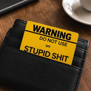 May include: A black leather wallet with a yellow card that reads "WARNING DO NOT USE on STUPID SHIT" in bold, black text. The card is partially inserted into a wallet pocket, with the text clearly visible. The background is a wooden surface.