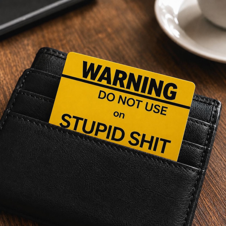 May include: A black leather wallet with a yellow card that reads "WARNING DO NOT USE on STUPID SHIT" in bold, black text. The card is partially inserted into a wallet pocket, with the text clearly visible. The background is a wooden surface.