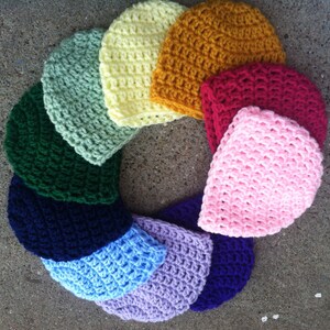 Pode incluir: Uma coleção de gorros de crochê coloridos e feitos à mão, dispostos em um padrão circular. Os gorros são em várias cores, incluindo verde, amarelo, laranja, vermelho, rosa, roxo e azul. Os chapéus são feitos de fio texturizado.