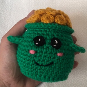 May include: A green crocheted plush toy with a yellow crocheted top, black eyes, and pink cheeks. The toy has a smiling mouth and is shaped like a pot.