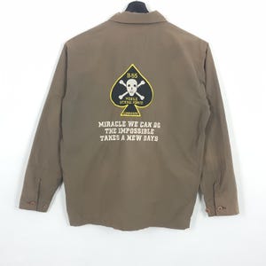 May include: A brown long-sleeve jacket featuring a black spade-shaped patch with a white skull and crossbones, yellow text reading B-55 MOBILE STRIKE FORCE COMMANDO, and white text below reading MIRACLE WE CAN DO THE IMPOSSIBLE TAKES A NEW DAYS.