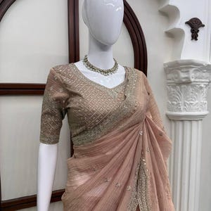 Puede incluir: Un sari de color melocot&oacute;n con una tela texturizada y plisada, con intrincados bordados plateados a lo largo de los bordes y una blusa a juego con escote en V. El conjunto se complementa con un collar de cuentas plateadas y peque&ntilde;as perlas.