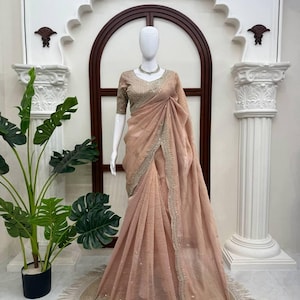 Puede incluir: Un sari de color melocot&oacute;n drapeado sobre un maniqu&iacute;, con una tela texturizada y un borde ancho adornado con bordados plateados y peque&ntilde;os espejos. La blusa a juego tiene mangas cortas y detalles plateados intrincados en toda la prenda.