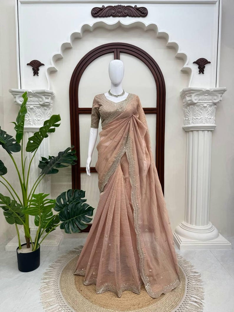Puede incluir: Un sari de color melocot&oacute;n drapeado sobre un maniqu&iacute;, con una tela texturizada y un borde ancho adornado con bordados plateados y peque&ntilde;os espejos. La blusa a juego tiene mangas cortas y detalles plateados intrincados en toda la prenda.