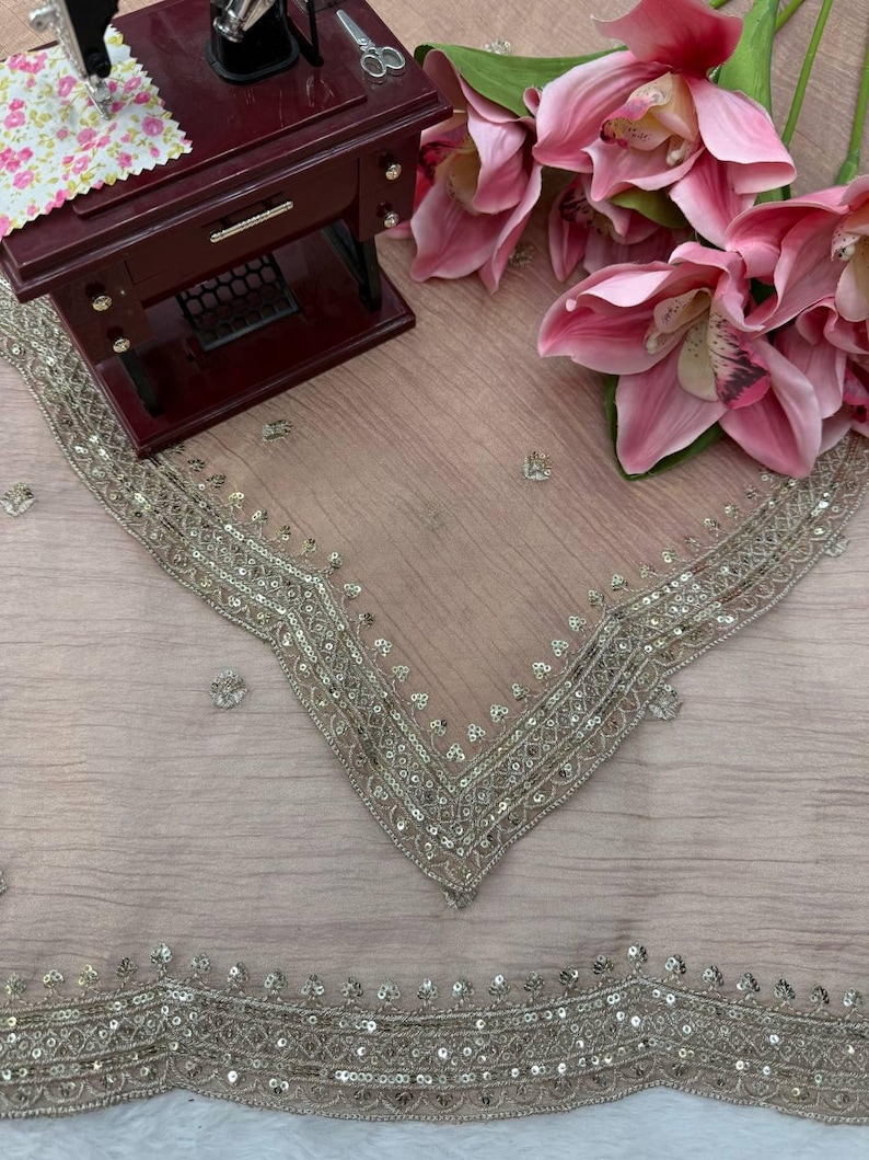 Puede incluir: Una tela transparente de color marr&oacute;n claro con un borde ancho e intrincado de lentejuelas plateadas y bordados met&aacute;licos. La tela est&aacute; drapeada junto a una m&aacute;quina de coser en miniatura de color granate y un ramo de grandes orqu&iacute;deas artificiales de color rosa vibrante.