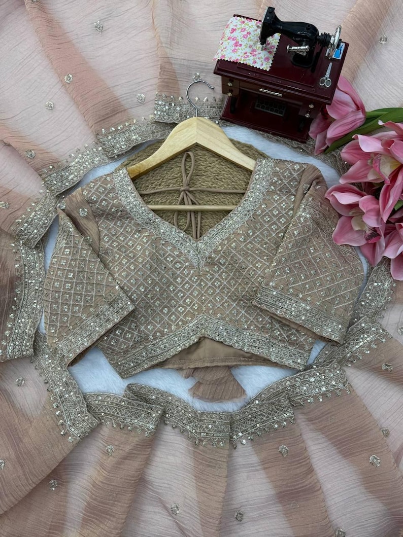Puede incluir: Un conjunto de blusa y falda en color melocot&oacute;n claro con intrincados bordados plateados en un patr&oacute;n de cuadr&iacute;cula de diamantes. La blusa tiene mangas cortas y cuello en V, mientras que la falda es de tela plisada transparente con detalles de lentejuelas plateadas.