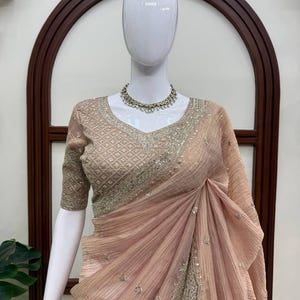 Puede incluir: Un sari de color melocot&oacute;n con tela texturizada y plisada con adornos de lentejuelas plateadas dispersas. La blusa a juego tiene un escote en V y un intrincado bordado plateado con un patr&oacute;n geom&eacute;trico, junto con un collar de cuentas plateado.