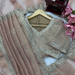 Puede incluir: Una prenda de tela transparente en color beige claro con intrincados bordados plateados y detalles de lentejuelas a lo largo de los bordes y el corpi&ntilde;o. La pieza incluye un escote en V y un drapeado plisado texturizado, acentuado por un peque&ntilde;o modelo de m&aacute;quina de coser.
