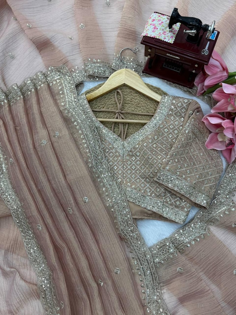 Puede incluir: Una prenda de tela transparente en color beige claro con intrincados bordados plateados y detalles de lentejuelas a lo largo de los bordes y el corpi&ntilde;o. La pieza incluye un escote en V y un drapeado plisado texturizado, acentuado por un peque&ntilde;o modelo de m&aacute;quina de coser.