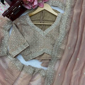 Puede incluir: Un conjunto de sari en color rosa empolvado que incluye una blusa a juego con intrincados bordados plateados en patr&oacute;n de rombos. La blusa tiene cuello en V y mangas cortas. La tela transparente del sari est&aacute; adornada con delicadas lentejuelas.