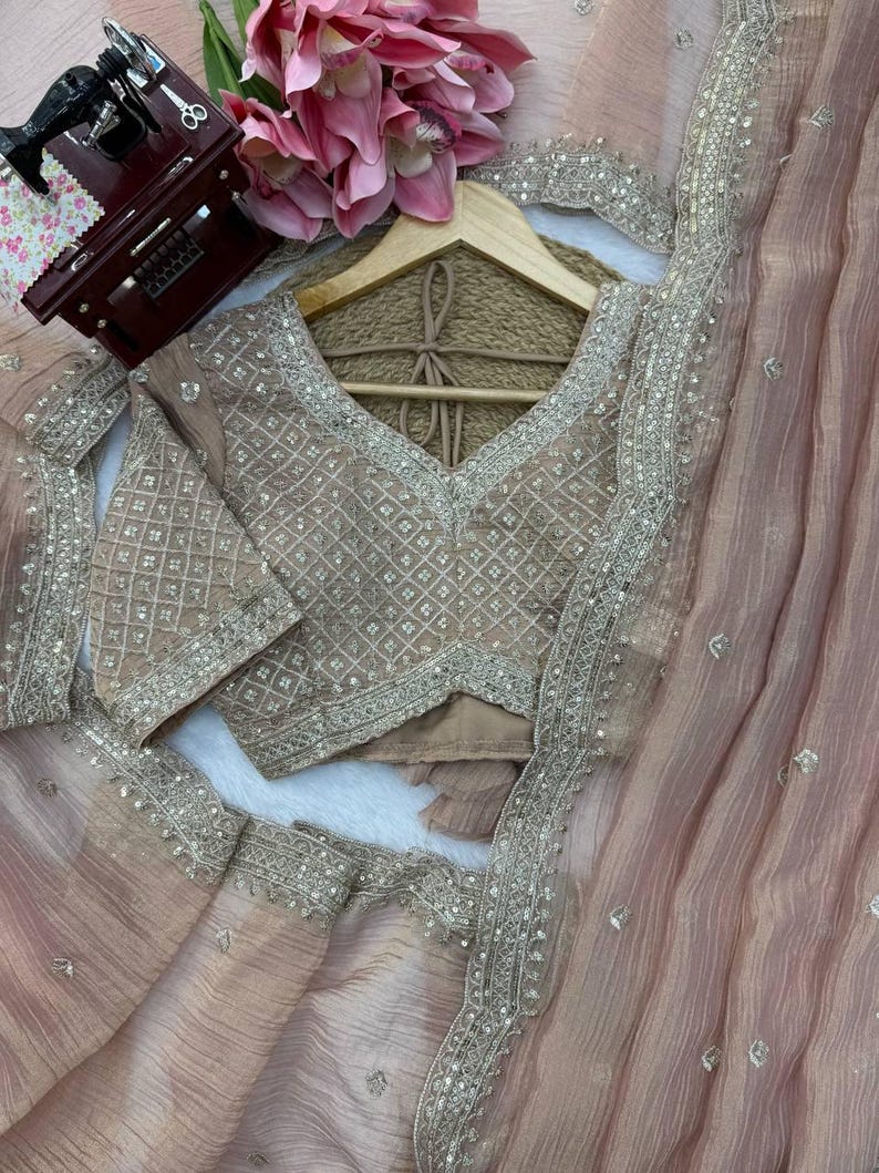 Puede incluir: Un conjunto de sari en color rosa empolvado que incluye una blusa a juego con intrincados bordados plateados en patr&oacute;n de rombos. La blusa tiene cuello en V y mangas cortas. La tela transparente del sari est&aacute; adornada con delicadas lentejuelas.