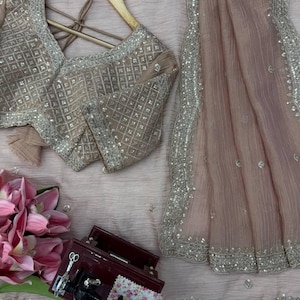 Puede incluir: Un conjunto rosa empolvado que incluye una blusa de manga corta con un dise&ntilde;o de lentejuelas plateadas en cuadr&iacute;cula y una tela drapeada transparente a juego. La blusa cuelga de una percha de madera junto a una peque&ntilde;a caja roja.