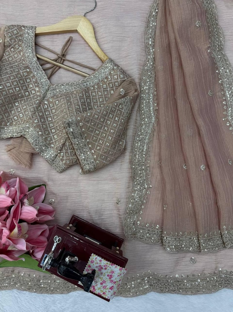 Puede incluir: Un conjunto rosa empolvado que incluye una blusa de manga corta con un dise&ntilde;o de lentejuelas plateadas en cuadr&iacute;cula y una tela drapeada transparente a juego. La blusa cuelga de una percha de madera junto a una peque&ntilde;a caja roja.