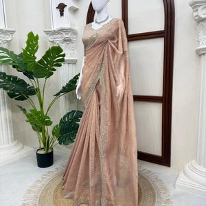 Puede incluir: Un sari de color melocot&oacute;n claro con un tejido texturizado de brillo sutil. La prenda incluye una blusa a juego y un borde ancho y ornamentado decorado con intrincados bordados plateados y peque&ntilde;os adornos reflectantes por toda la pieza.