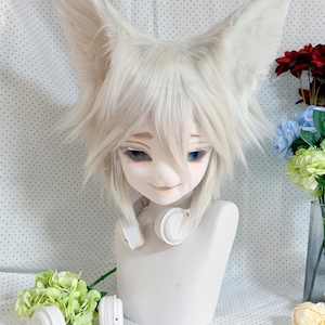 May include: A white anthropomorphic animal mask featuring large, pointed, furry ears and a layered, shoulder-length white wig. The mask has stylized blue eyes, a small nose, and a neutral expression, worn with white over-ear headphones.